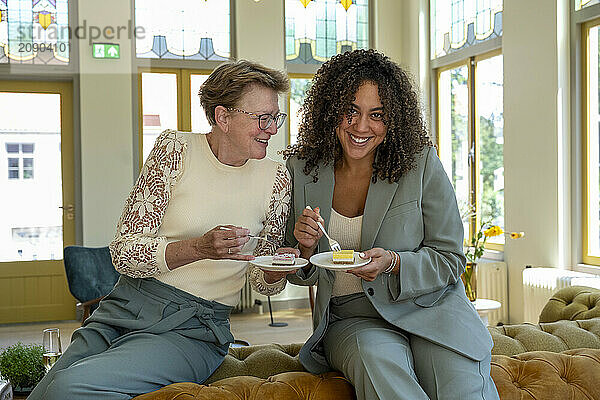 Two women enjoying tea and conversation on a sofa in a well-lit room with stained glass windows.