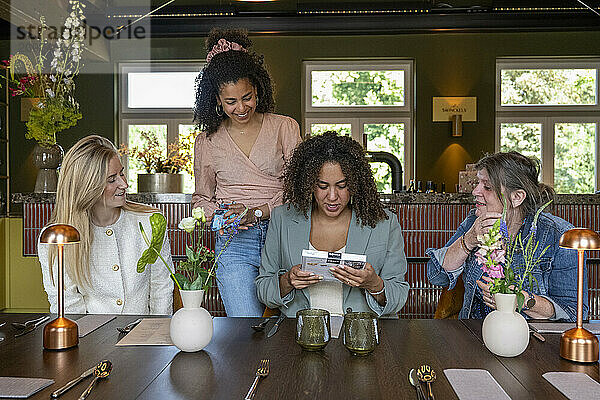 Four women enjoying a moment together around a table with one showing a card to others  amidst elegant interior decor.