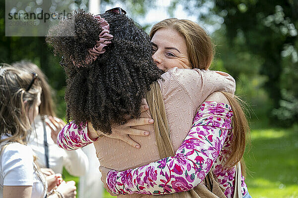 Two women embracing in a warm hug outdoors with people in the background.