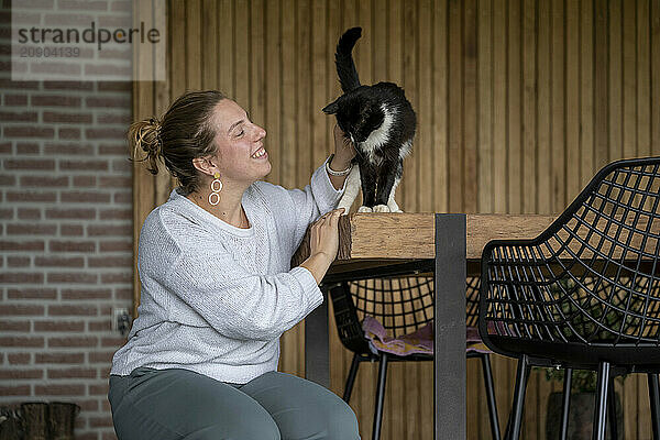 Smiling woman enjoys time with a black and white cat at a wooden table.