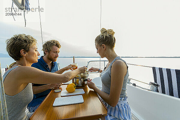 Three people enjoy a meal together on the deck of a sailboat with a serene water backdrop during sunset.