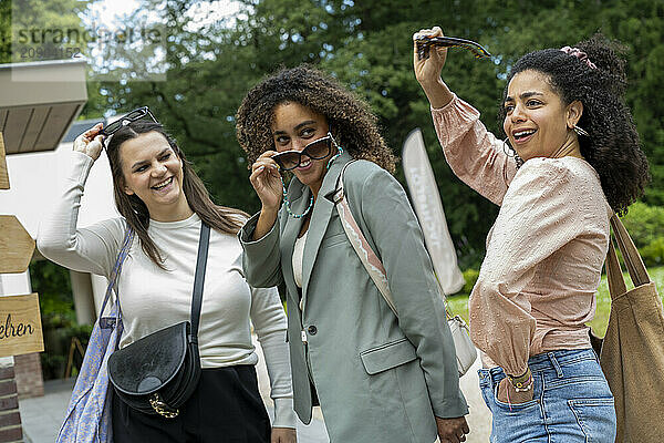 Three women smiling and trying on sunglasses at an outdoor market.