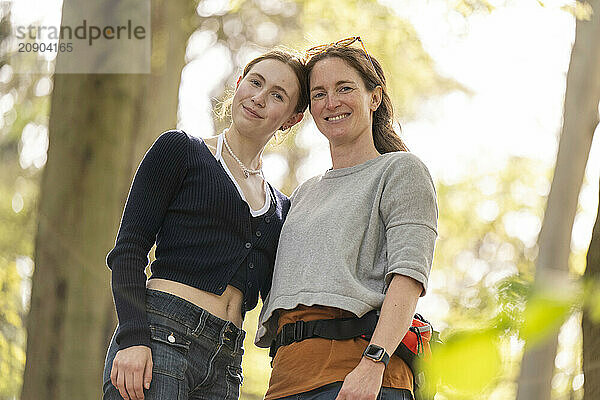 Two smiling women embracing outdoors with sunlight filtering through trees in the background.