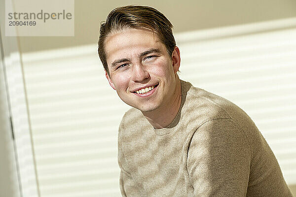 Smiling young man in a beige sweater posing indoors with a bright background.