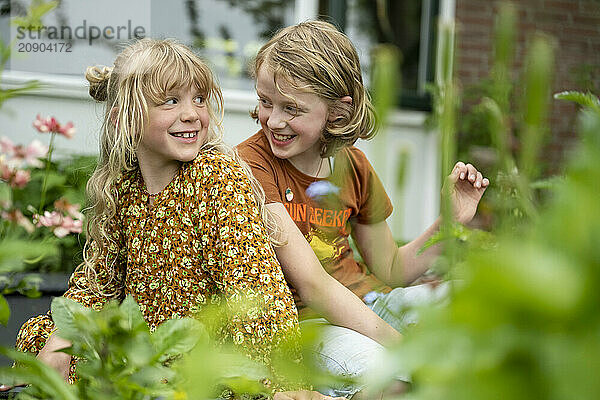 Two smiling girls sitting together in a garden surrounded by plants and flowers on a sunny day.