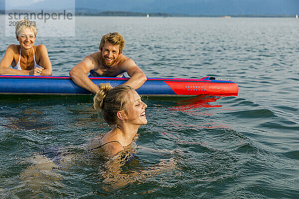 Three friends enjoy a day at the lake with one woman swimming in the foreground and two people on a paddleboard in the background.