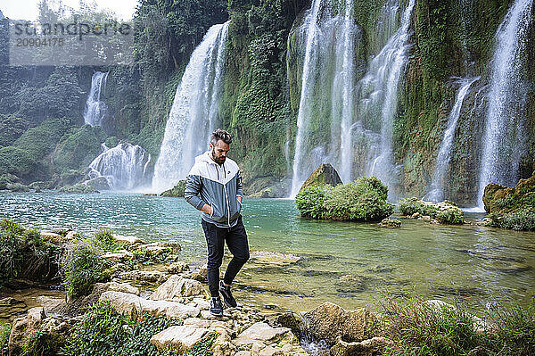 Man hiking on rocky terrain with a majestic waterfall and lush foliage in the background.