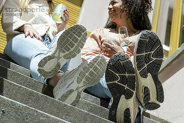 Two women laughing and enjoying drinks while sitting on outdoor steps  casually dressed with the soles of their shoes prominently displayed.