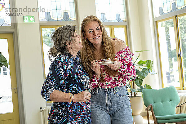 Smiling older woman and young woman enjoying time together  holding a plate with a dessert in a brightly lit room with elegant windows.