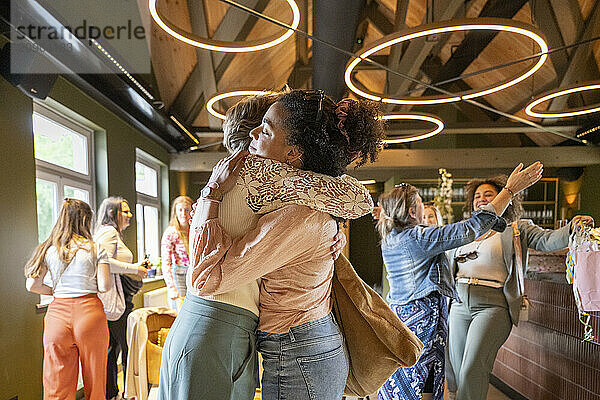 Two women embracing joyfully in a warm hug at a vibrant indoor gathering with other people chatting in the background.