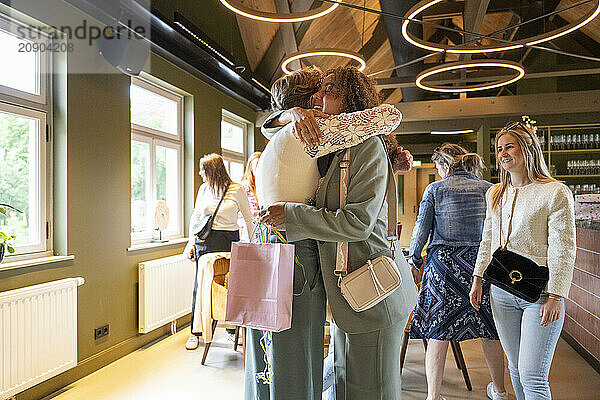 Two friends hug warmly in a cozy cafe while another woman looks on with a smile.