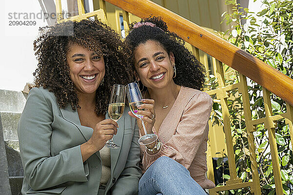 Two women with curly hair smiling and holding glasses of wine on an outdoor staircase.