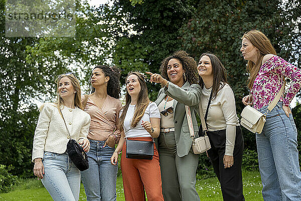 A group of six smiling women enjoying time together outdoors  with one pointing at something in the distance.