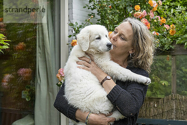 Woman affectionately kisses a white puppy she is holding in a garden.