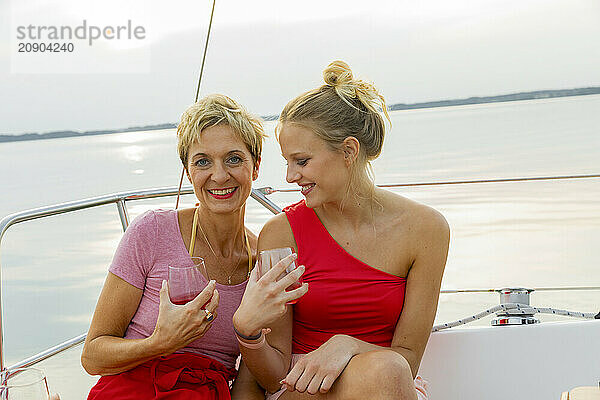 Two women smiling and holding glasses on a boat with a lake view at sunset.