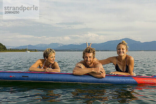 Three friends smiling on a blue kayak in a lake with mountains in the background.