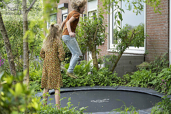 Children playing on a garden trampoline with a house and greenery in the background.