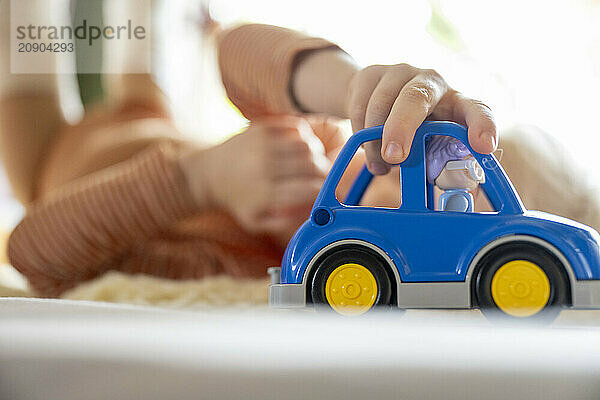 Child playing with a blue toy car on a soft surface with a focus on the car and the child in the blurred background.