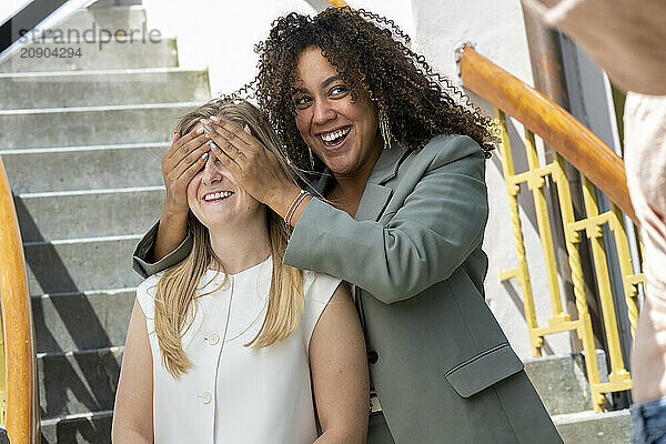 A joyful woman with curly hair surprises another woman by covering her eyes with her hands on a staircase outdoors.