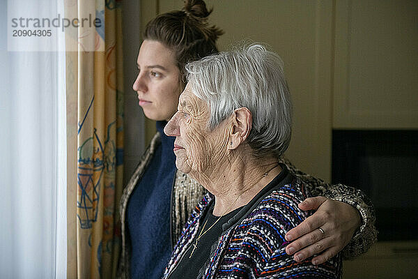 A caring young woman gently places her hand on an elderly woman's shoulder as they both look out the window thoughtfully.