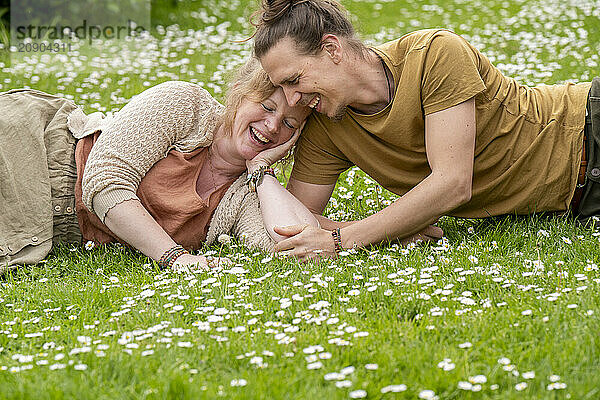 A smiling man and woman lie close together on a grassy field dotted with white flowers  enjoying a tender moment.