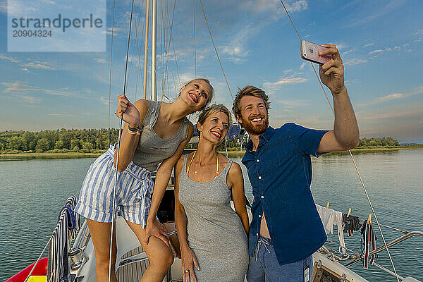 Three friends take a selfie on a sailboat with a scenic lake and clear skies in the background.