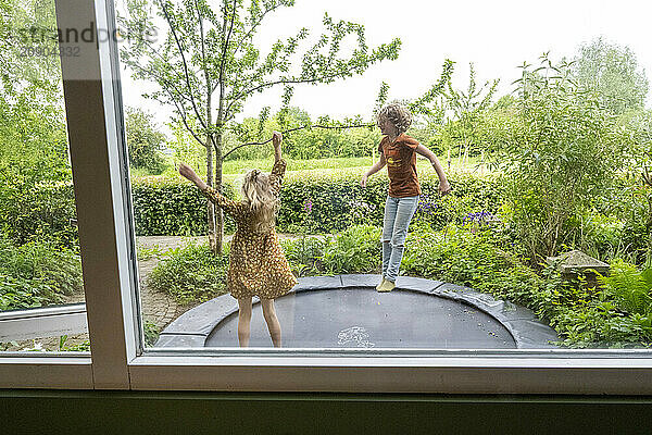 Two children are joyfully jumping on an outdoor trampoline in a lush garden  viewed from inside through an open window.