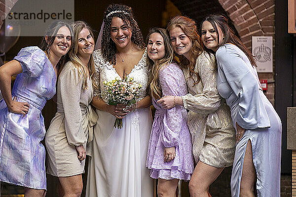 A smiling bride in a white dress surrounded by five bridesmaids in pastel dresses  posing together outdoors.