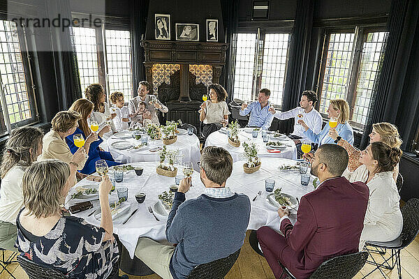 A group of people enjoying a festive gathering around a dinner table decorated with flowers and place settings  engaging in conversation and toasting with drinks.