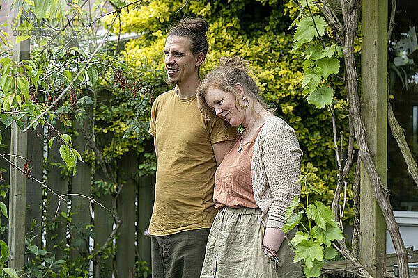 A man and woman smile gently while leaning against each other in a lush garden.