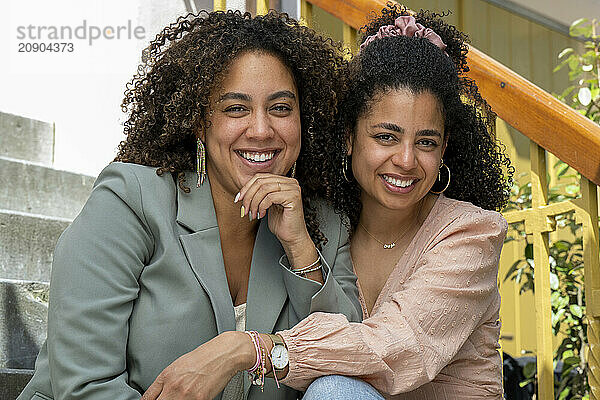 Two smiling women sitting together on steps  one in a green blazer  the other in a pink jacket  both appearing happy and relaxed.