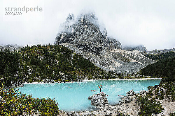 A person stands on a rock with arms raised in triumph in front of a stunning turquoise alpine lake with a majestic mountain shrouded in mist in the background.