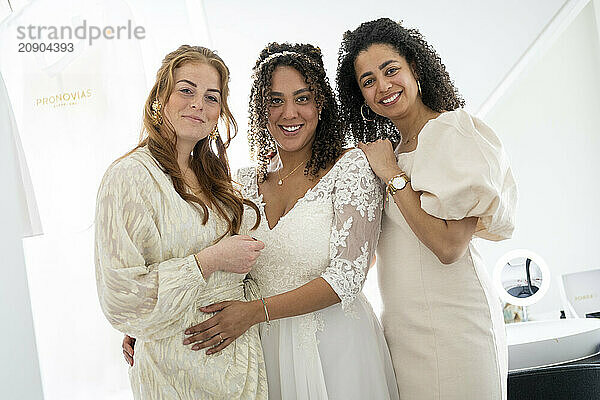 Three smiling women dressed elegantly  one in a white bridal gown  posing together indoors.