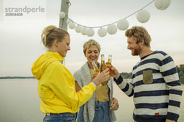 Three friends enjoying a beachside party with festive lights and a toast at sunset.