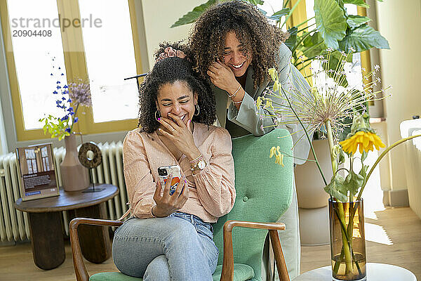 Two women share a joyful moment while looking at a smartphone together surrounded by houseplants.