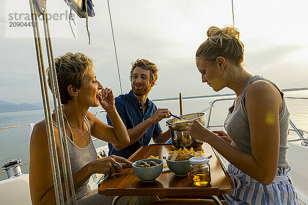 Friends enjoy a meal on a sailboat at sunset with a scenic water landscape in the background.