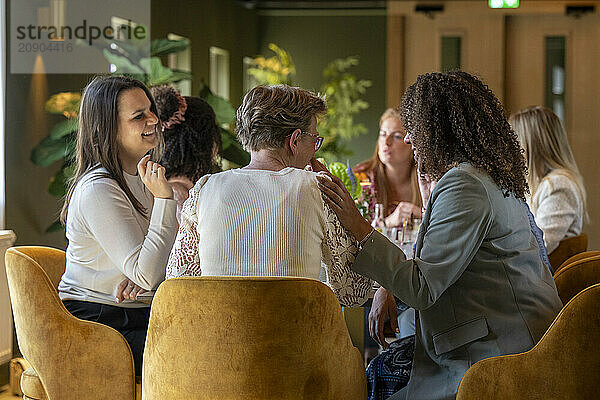 A group of women engaged in conversation around a table in a cozy restaurant setting with warm lighting.