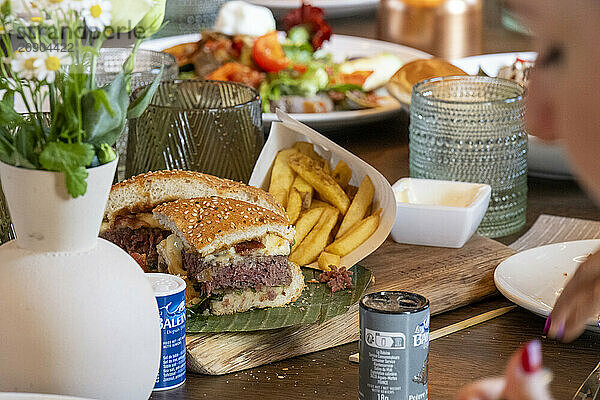 Close-up of a delicious burger cut in half with fries and assorted condiments on a wooden table  with an out-of-focus person in the foreground.