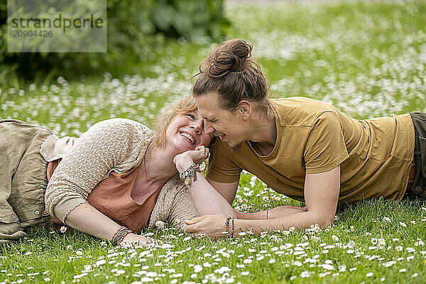 Two women are lying on a grassy field with white flowers  smiling and looking at each other affectionately.