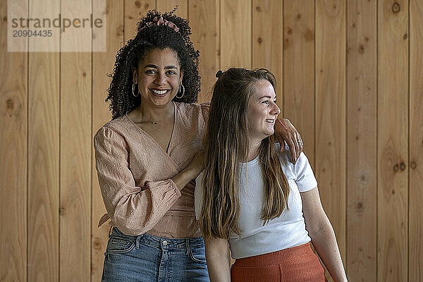 Two smiling women standing close together against a wooden wall background  looking to their right.