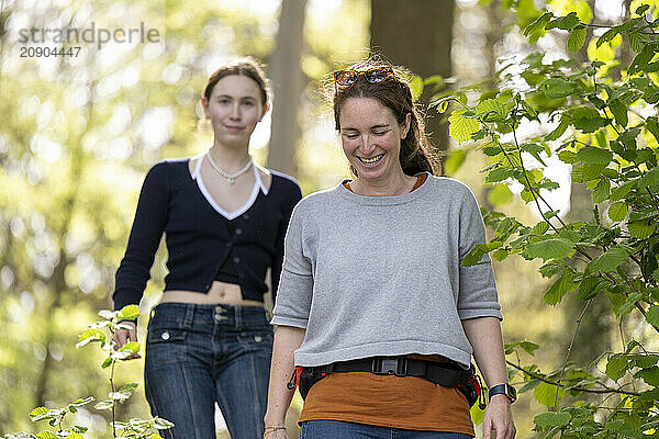 Two women walking through a sun-dappled forest  smiling and enjoying a casual hike on a bright day.