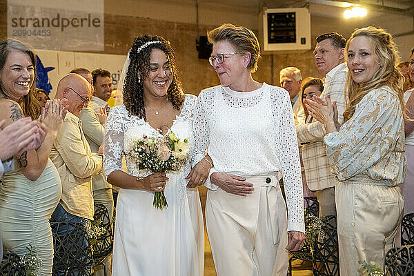 A joyful bride holding a bouquet walks through a clapping crowd accompanied by an older woman in a festive indoor setting.