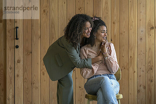 Two women share a joyful moment  with one standing and embracing the other who is seated  both smiling against a wooden panel background.