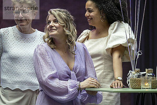 Three women in semi-formal attire happily engaging at a social event  standing behind a table with decorative items.