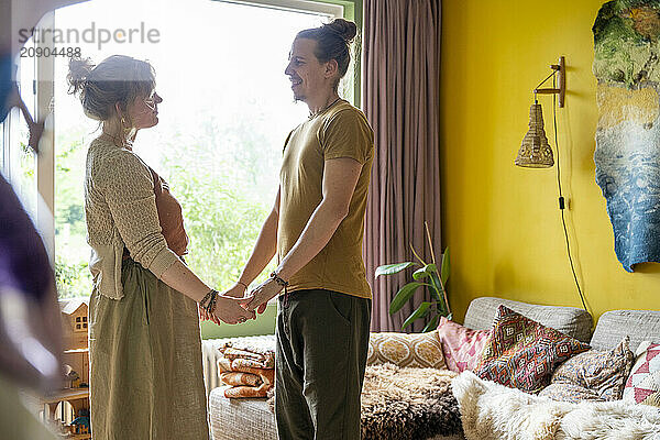 A couple holds hands lovingly while standing in a cozy living room decorated with a world map  plants  and eclectic furniture.