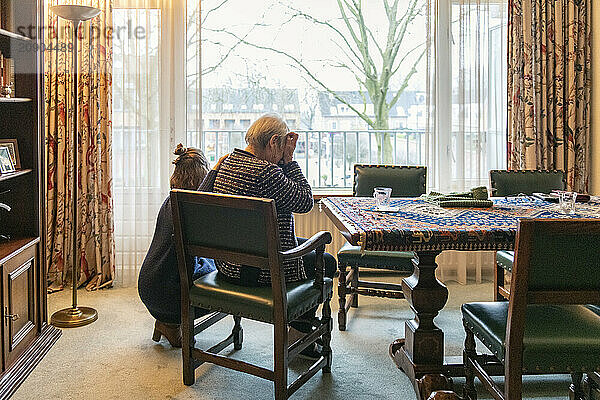 An elderly person sitting at a dining table in a cozy room  gazing out of a large window with a child playing nearby.