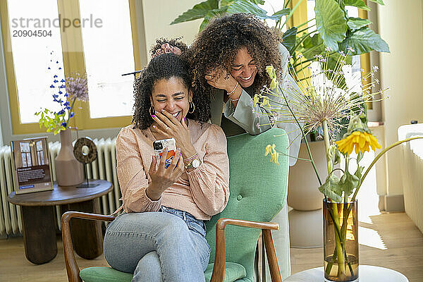 Two women smiling and sharing a moment with a smartphone in a sunny room with houseplants.