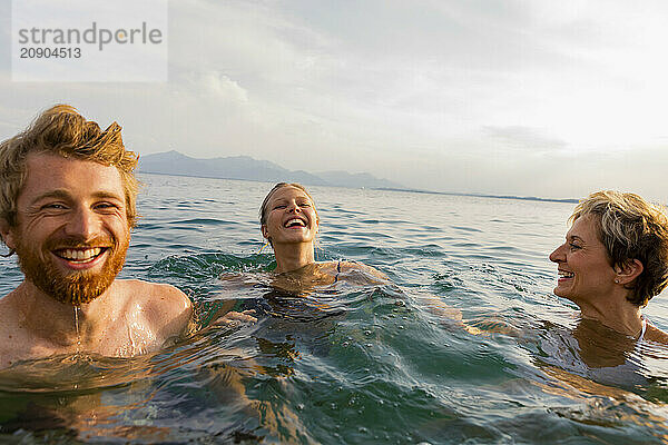 Three joyful people swimming and laughing together in the sea with a clear sky in the background.