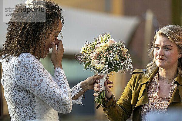 Emotional bride in lace dress wiping away tears while holding a bouquet as she shares a heartfelt moment with a woman in a jacket.