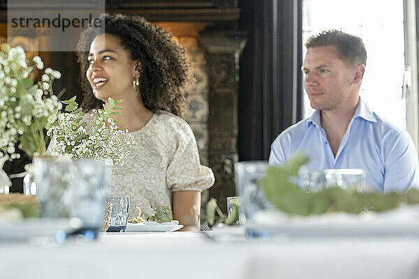 A woman with curly hair smiling and looking away at an event  with a man sitting beside her appearing distracted  both at a table elegantly set with white flowers.